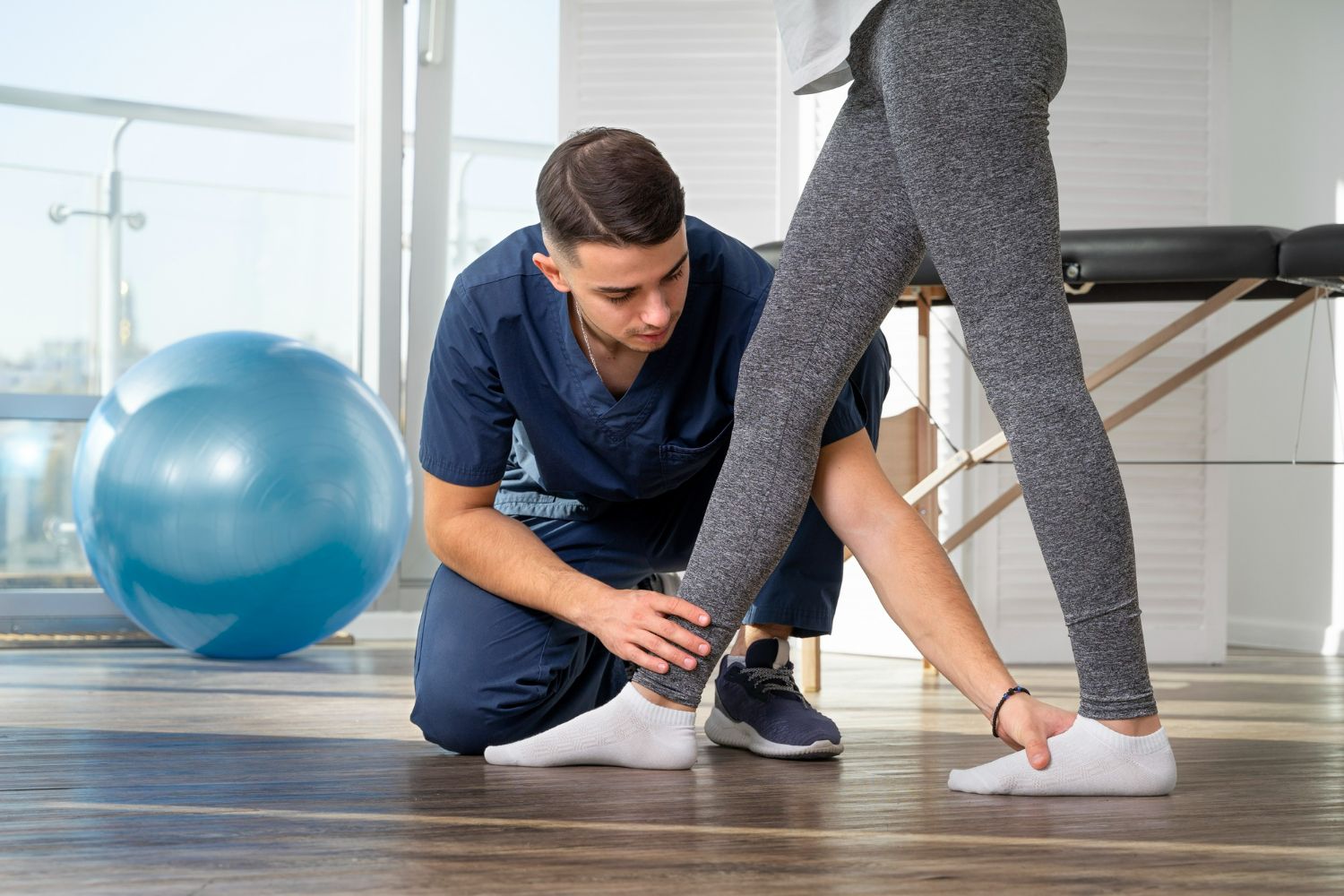 Physical therapist examining patient's ankle in a clinic. Blue ball, wooden floor, and a massage table are in the background.