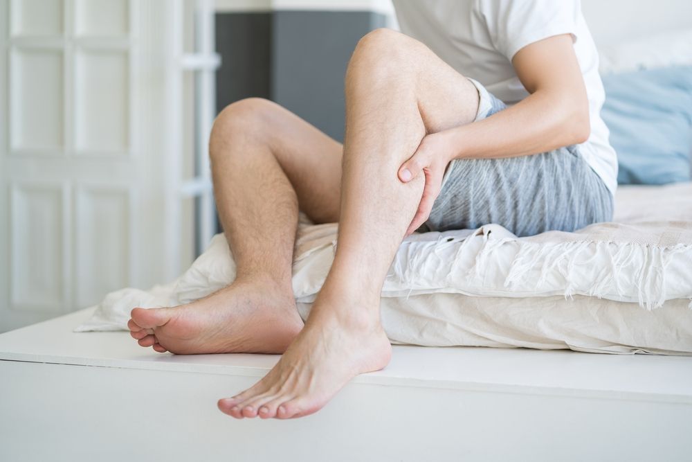 Man sitting on a bed, holding his calf as if in pain. Bright room, natural light.