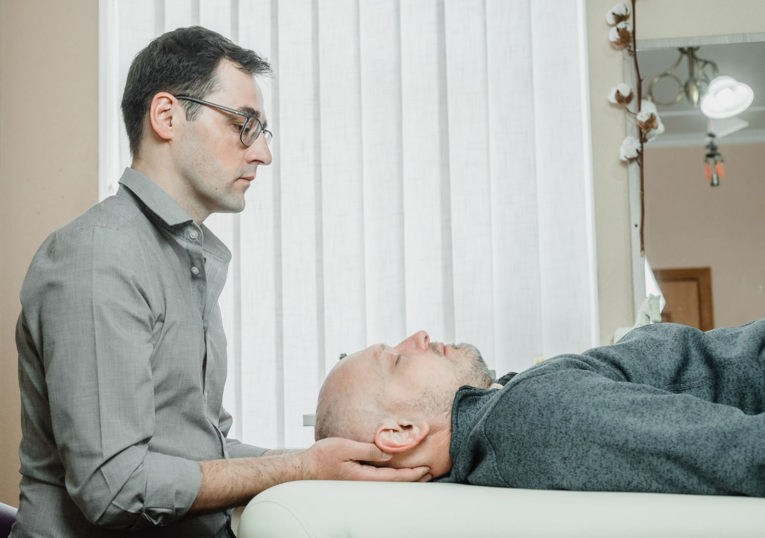 Man in glasses performing neck manipulation on a patient lying on a table. Light room.