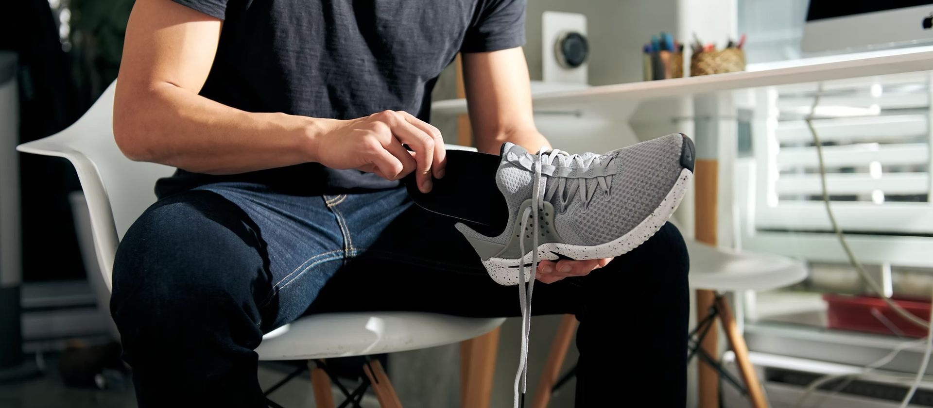 Person tying a gray running shoe while seated in a chair, with a white chair and bright room in the background