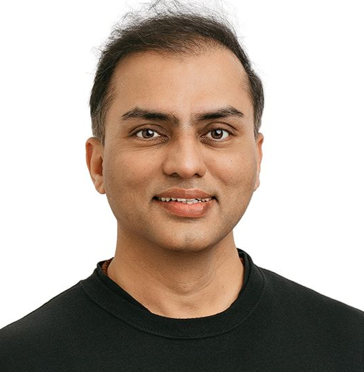 A smiling person wearing a black sweater and grey pants, posing with hands in pockets against a white background.