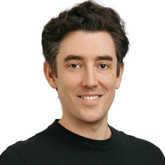 A person with short, dark curly hair, wearing a black t-shirt, smiling at the camera with one hand in their pocket.