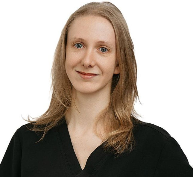A person with long blonde hair wearing a black V-neck scrub top, posing with one hand on their hip against a white background.