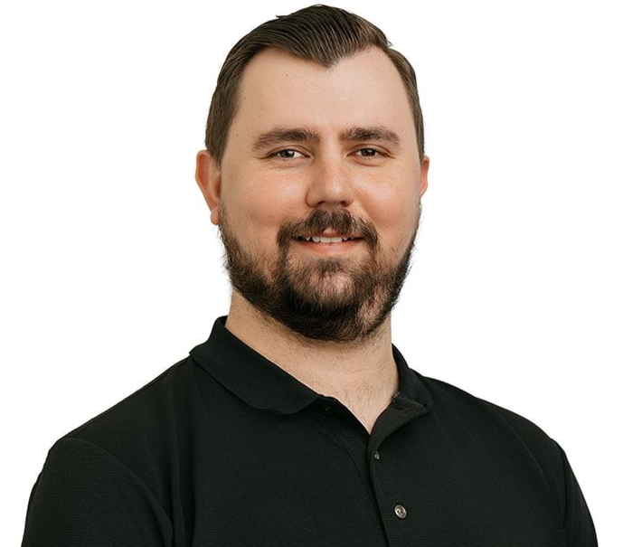 A person with a beard and tattoos on their arm, wearing a black polo shirt, stands against a white background.