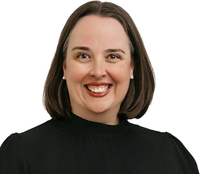 A person with shoulder-length brown hair, wearing a black long-sleeved textured top, smiling with hands clasped together.