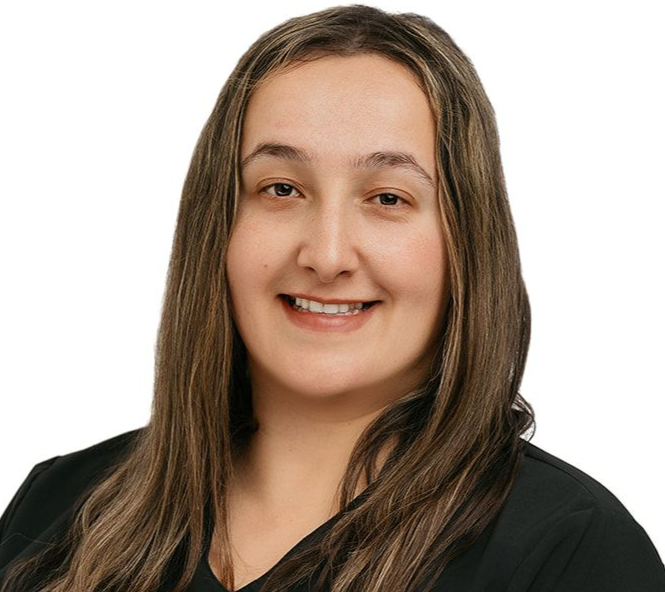 A person with long brown hair wearing a black V-neck scrub top, smiling and standing with their hands clasped.