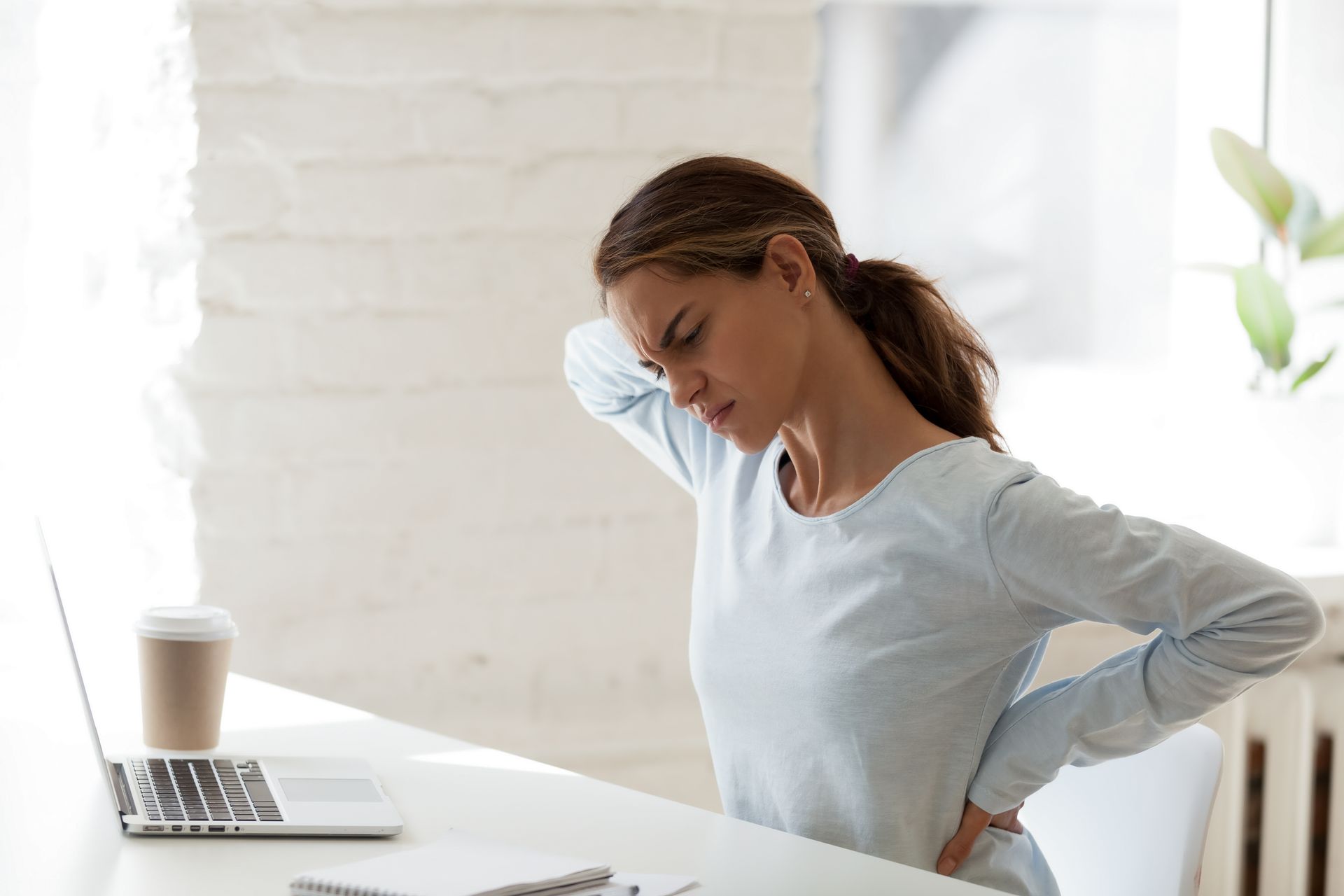 Woman holding her lower back while sitting at a desk with a laptop and coffee, looking strained