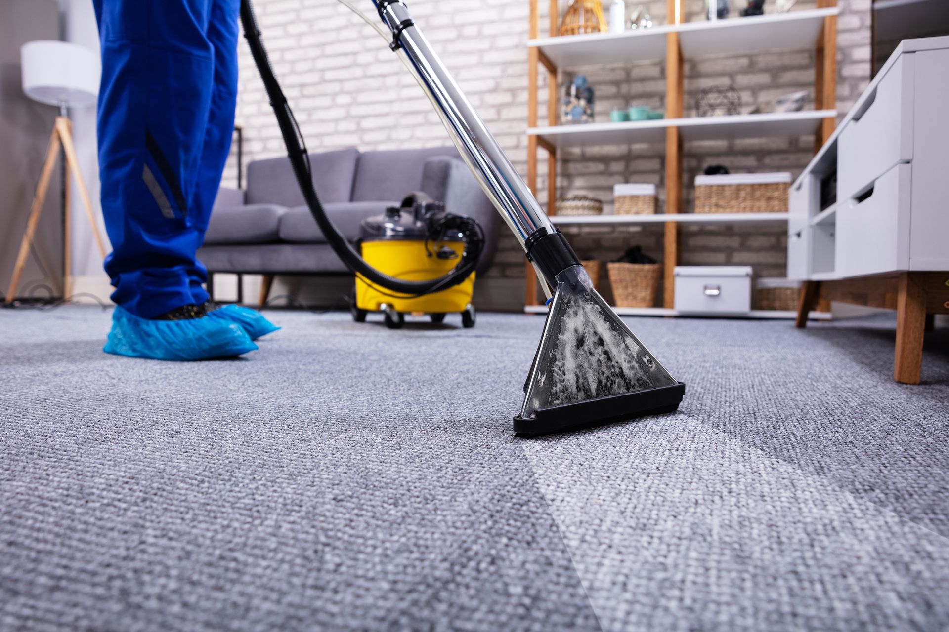 A carpet cleaner, wearing a blue uniform, uses a vacuum cleaner in a living room.