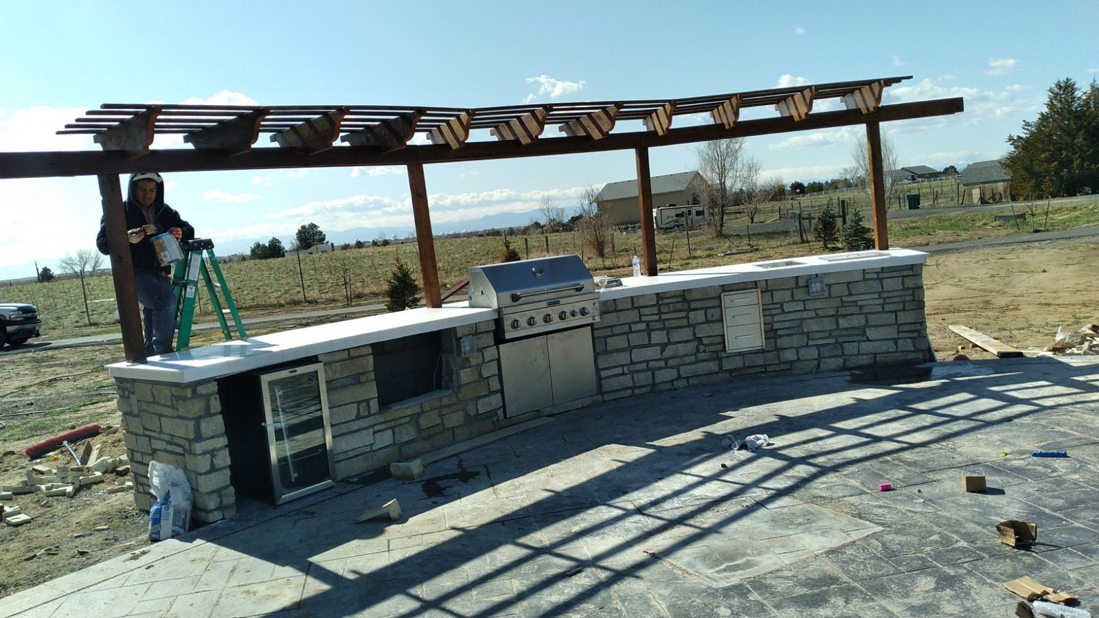 A man is standing on a ladder next to an outdoor kitchen under a pergola.