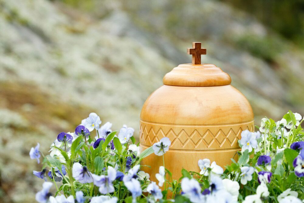 Wooden urn with a cross on top, among purple and white flowers.