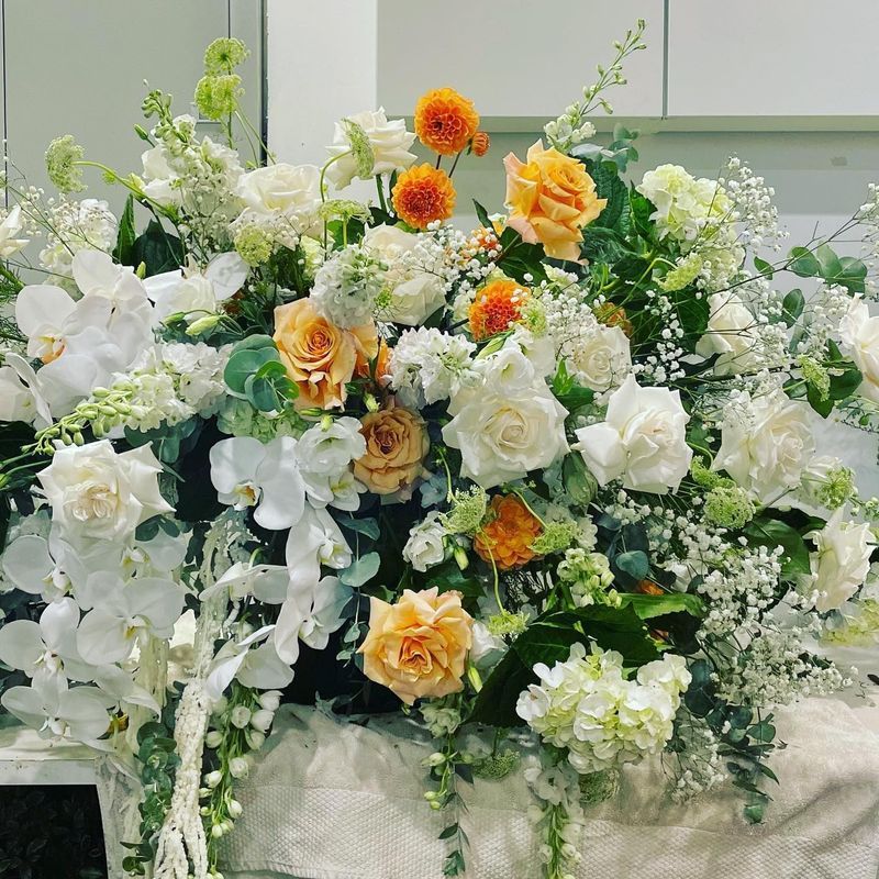 Floral arrangement: white orchids, roses, and orange flowers, with greenery.