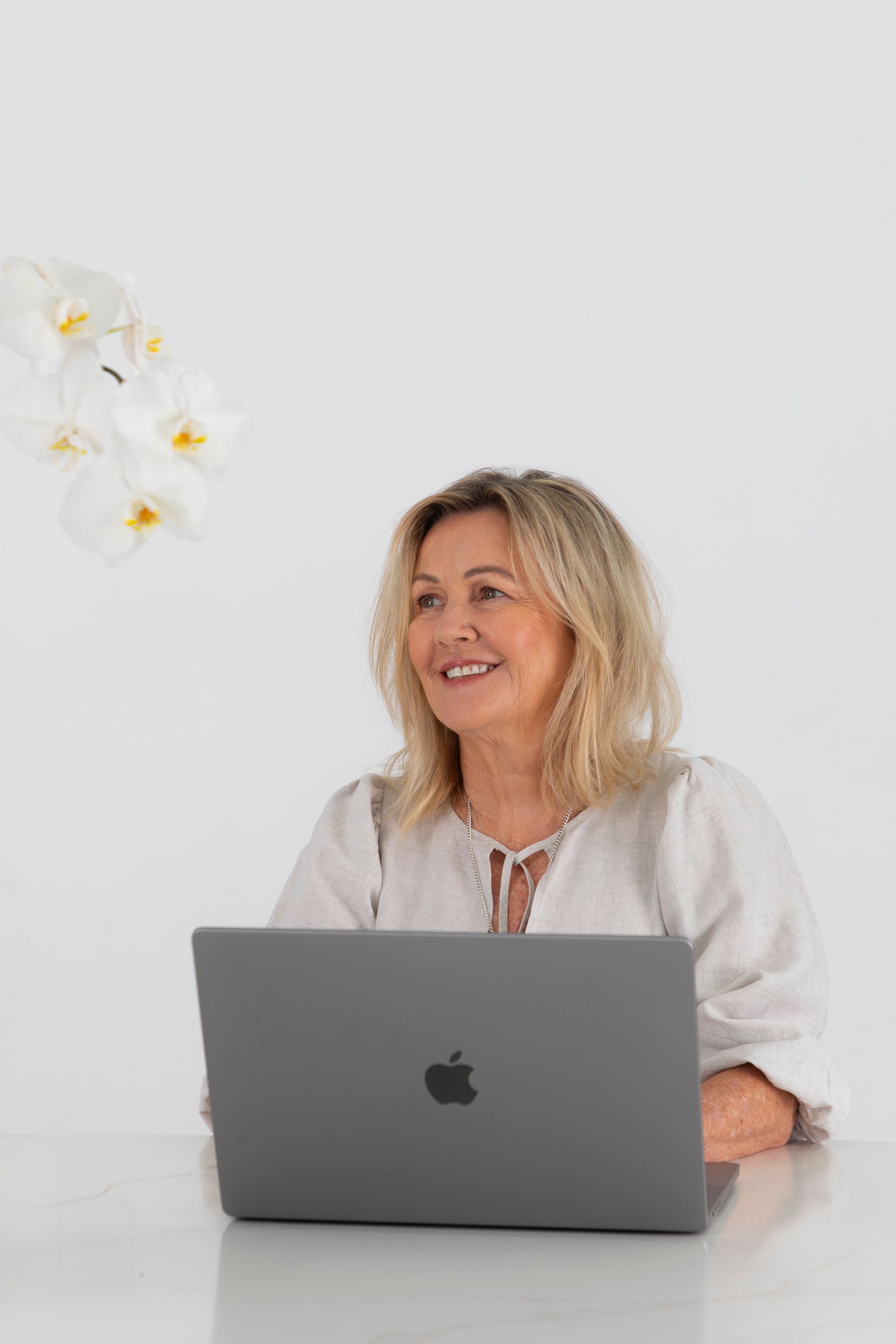 Blonde woman smiling, using a gray laptop at a white table, with orchids against a white backdrop.