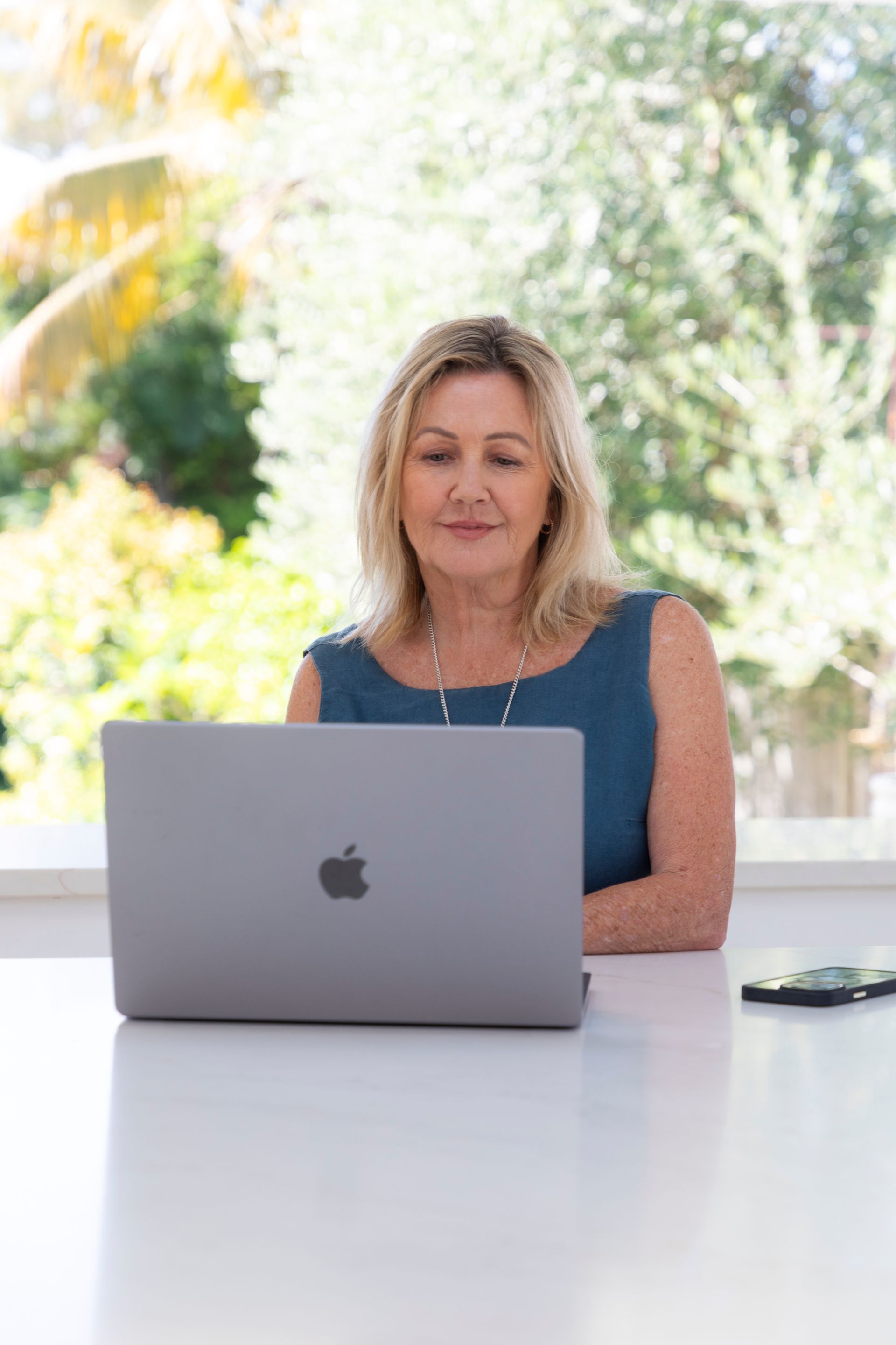 Woman with blonde hair, wearing a blue dress, sitting at a table using a laptop, with trees in the background.