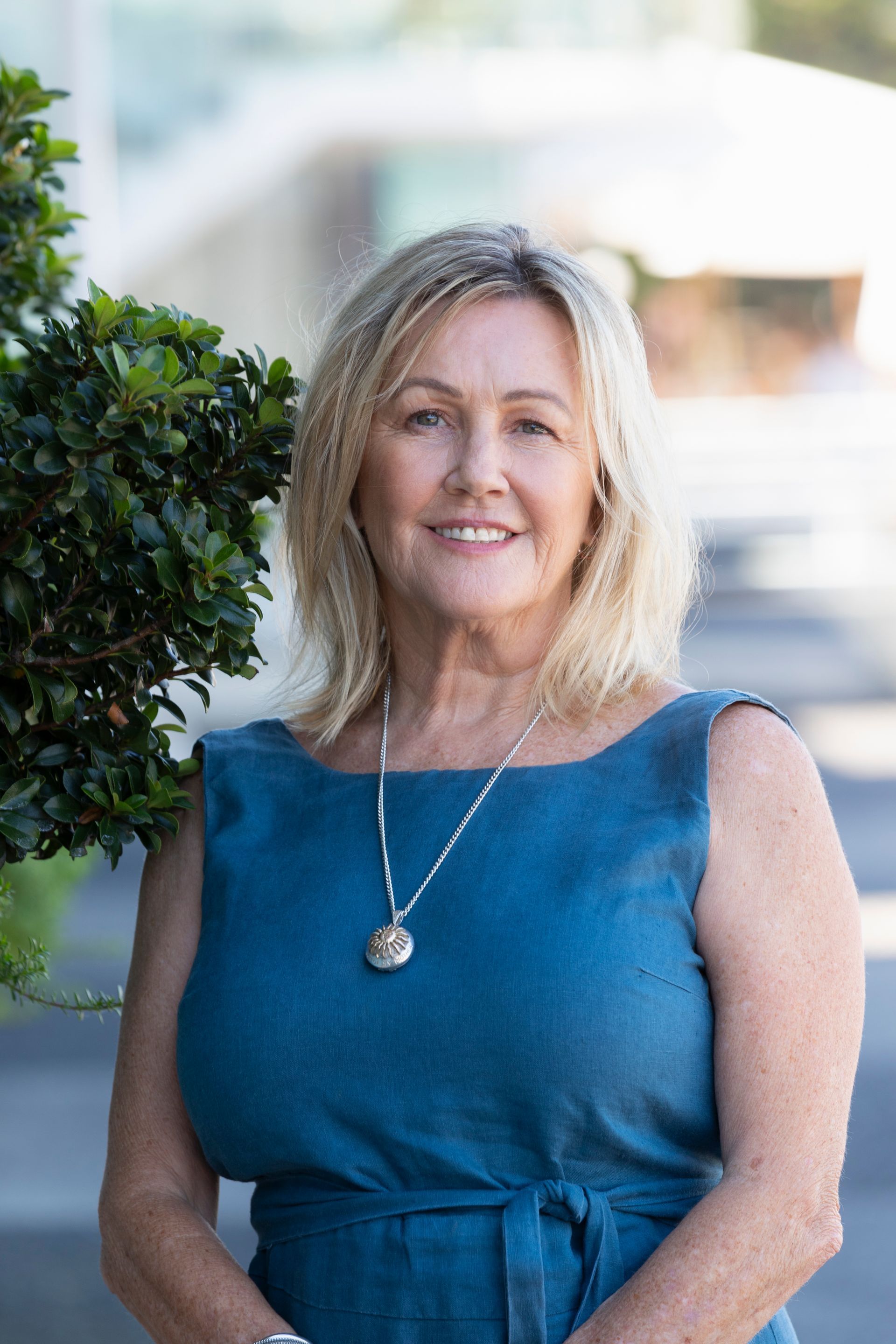 Blond woman smiling, wearing blue dress and necklace, standing near green foliage.