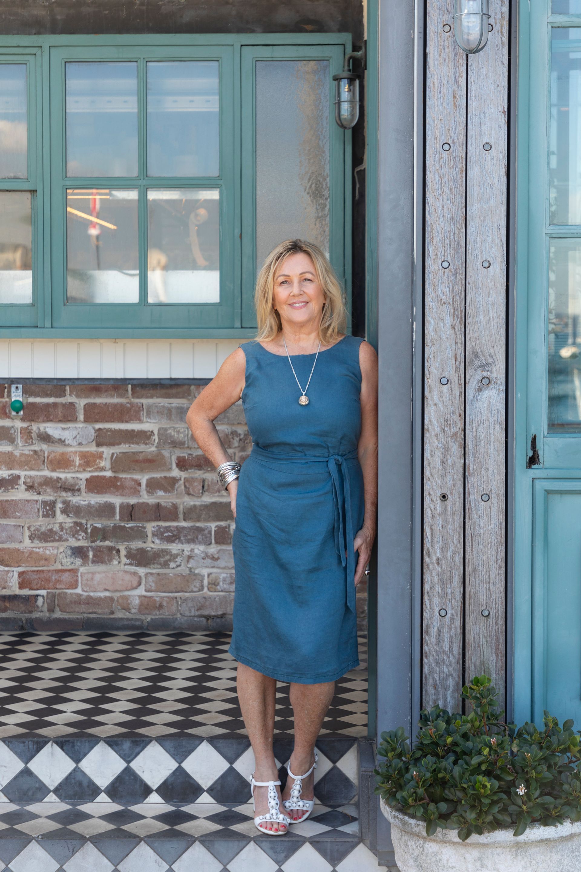 Woman in blue dress smiles, poses by brick building with teal accents, checkerboard steps.
