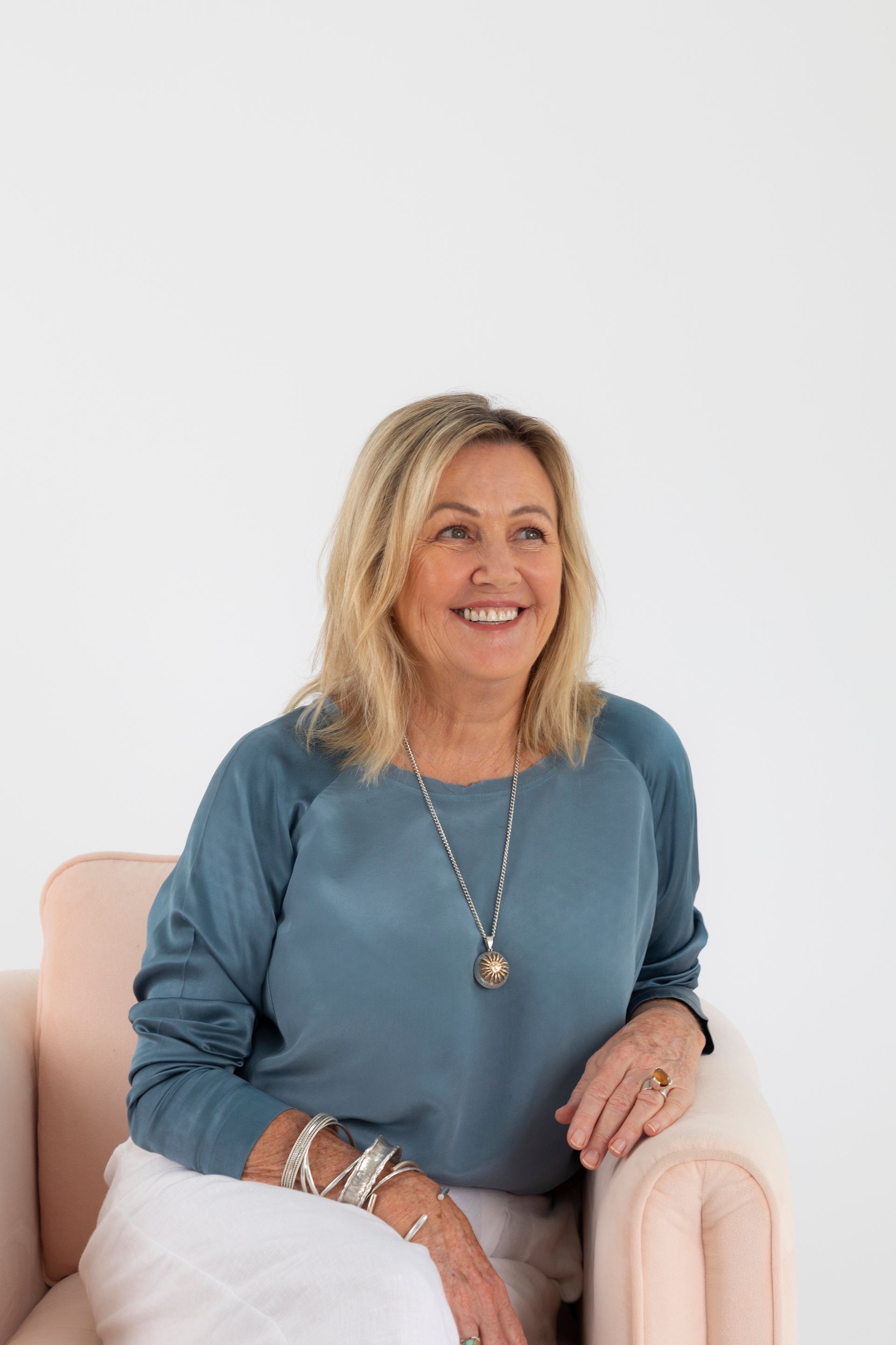 Woman with blonde hair smiling, sitting in a pink chair, wearing blue top and white pants.