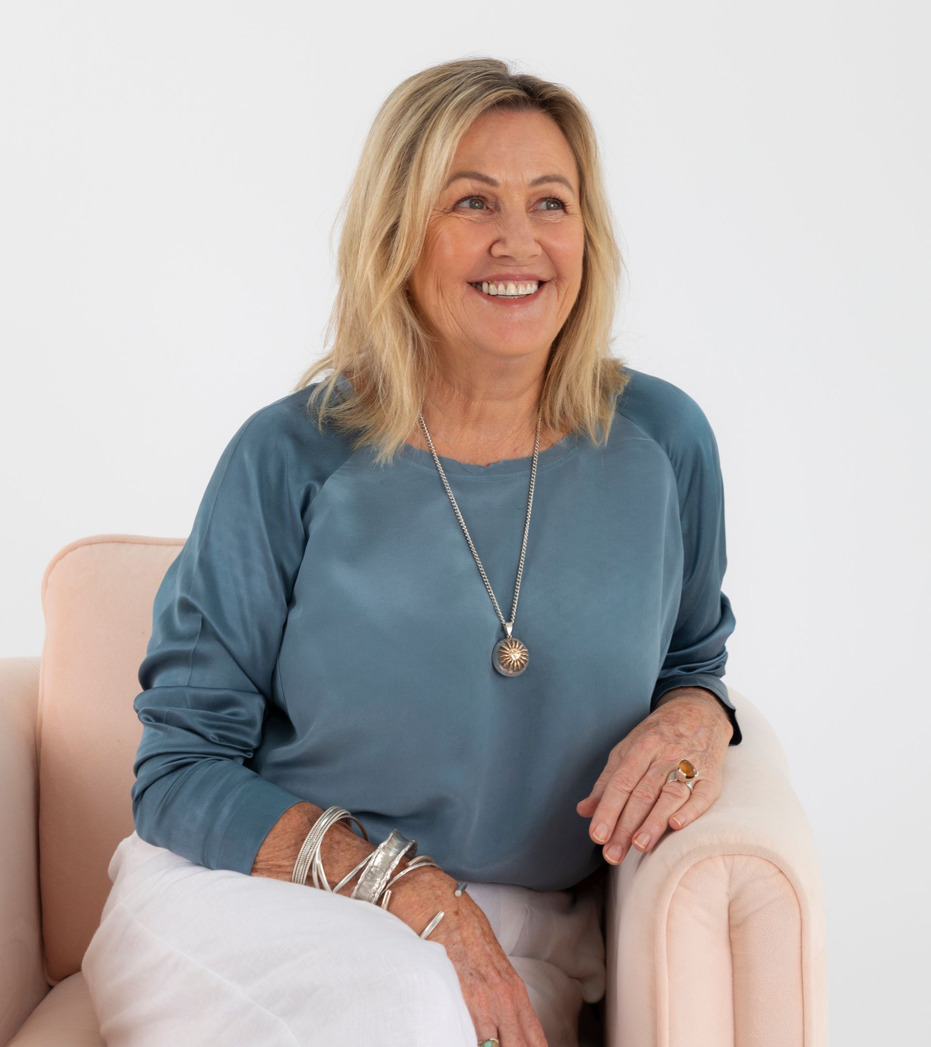 Blonde woman smiling, seated in pink chair, wearing blue top, white pants, jewelry, looking right.