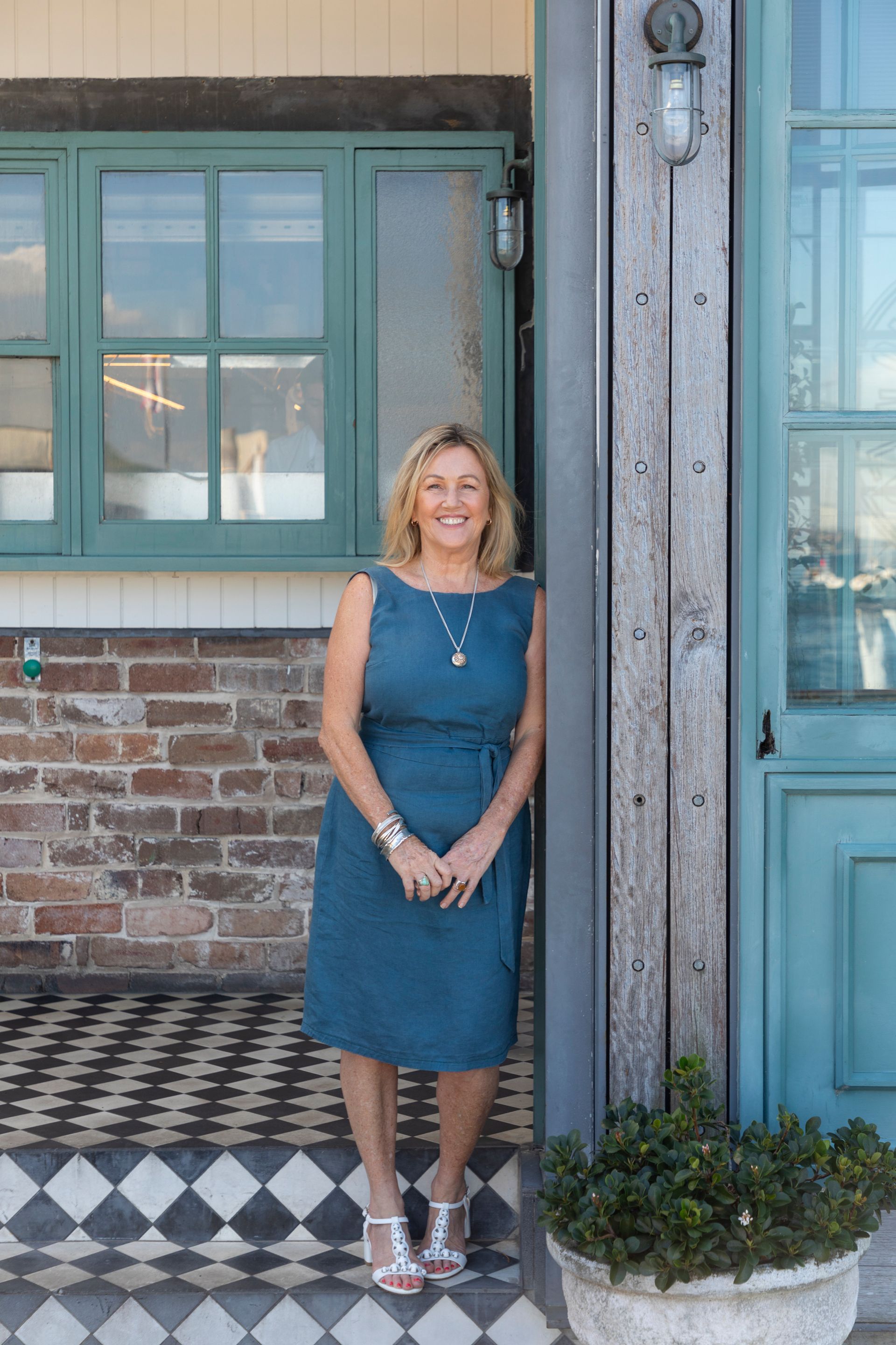 Woman in blue dress stands by a teal door and window with a brick wall in an outdoor setting.