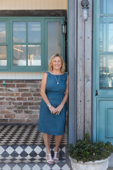 Woman in blue dress standing by a teal door and window, brick wall, and tiled steps.