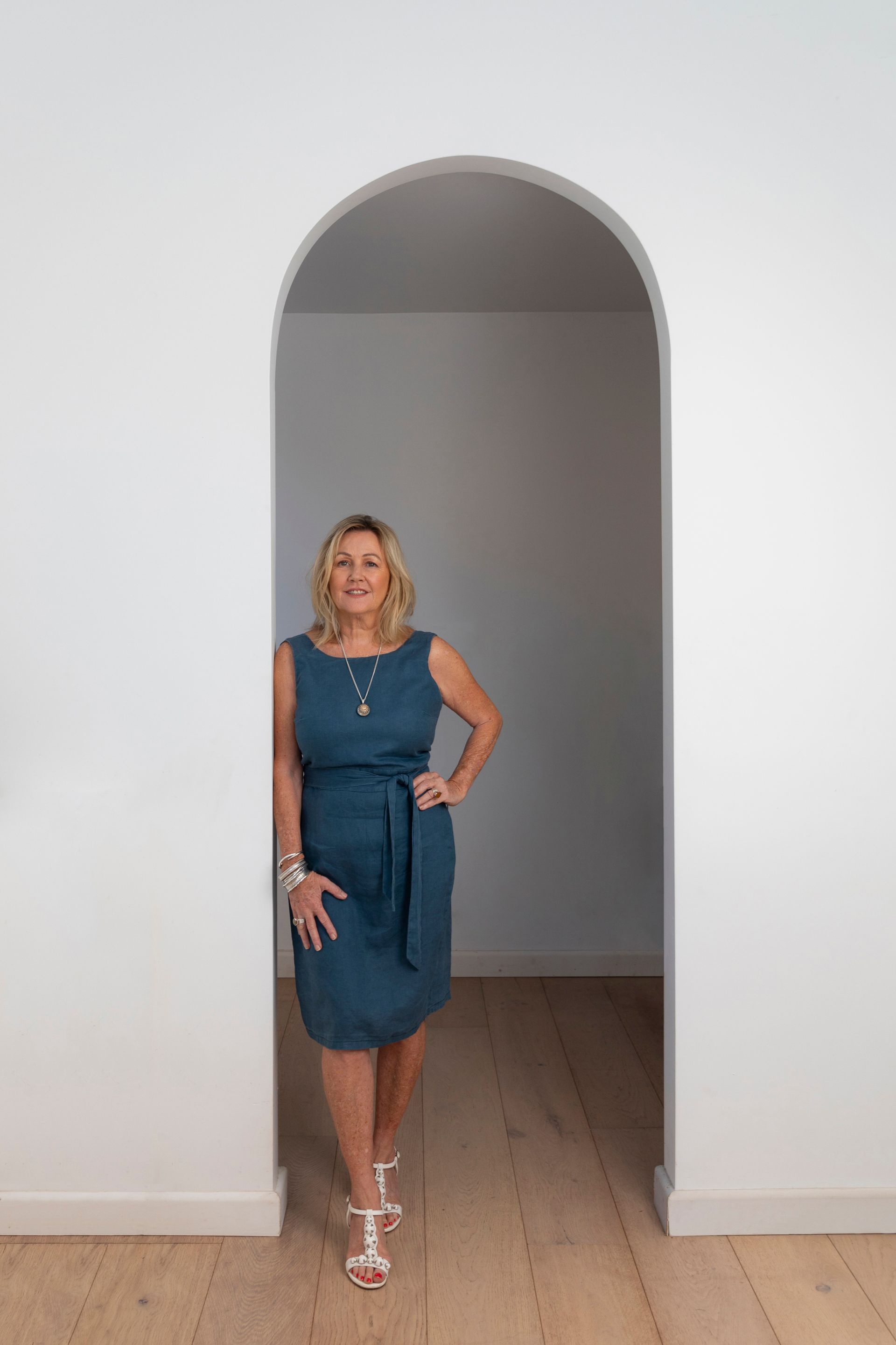 Woman in teal dress stands in archway, smiling, hand on hip. Blonde hair, white heels.