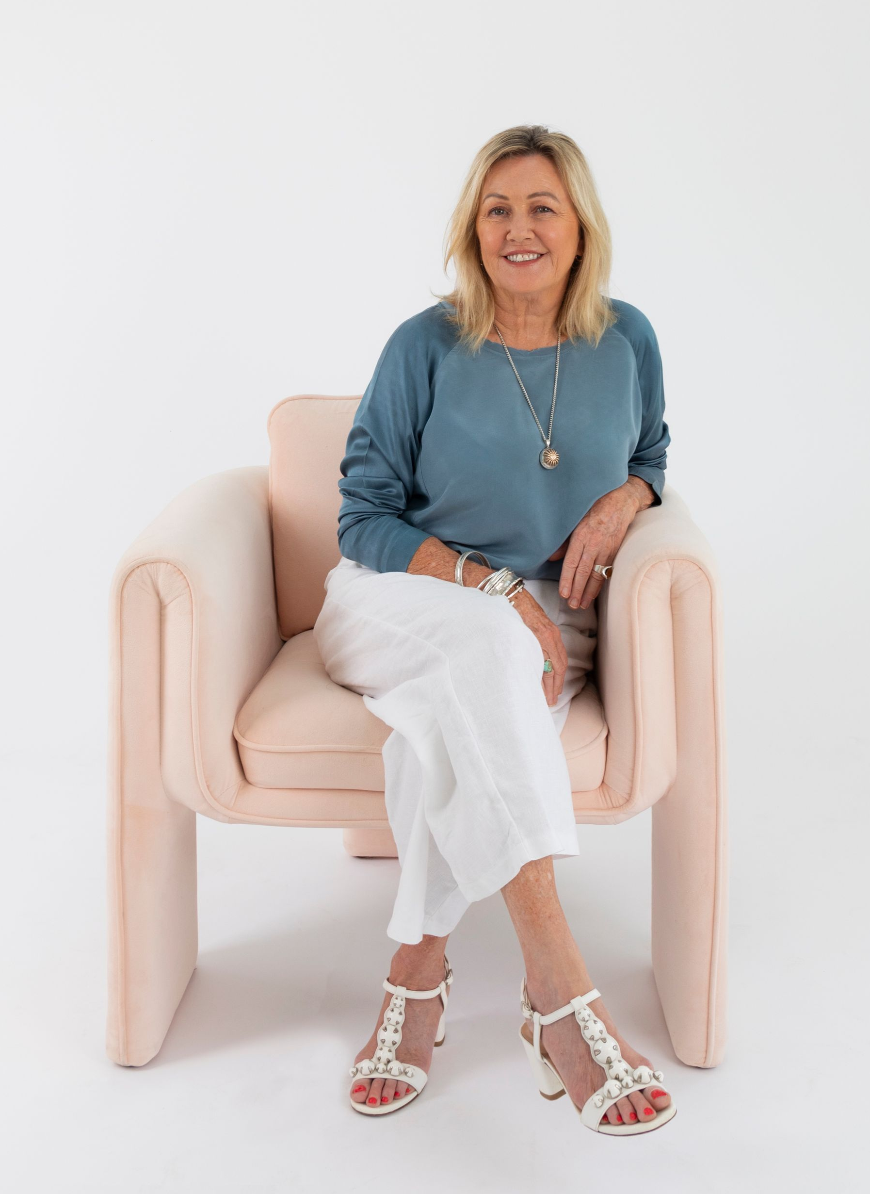 Woman with blonde hair, smiling, sitting in a pink chair, wearing blue top, white pants, and sandals.