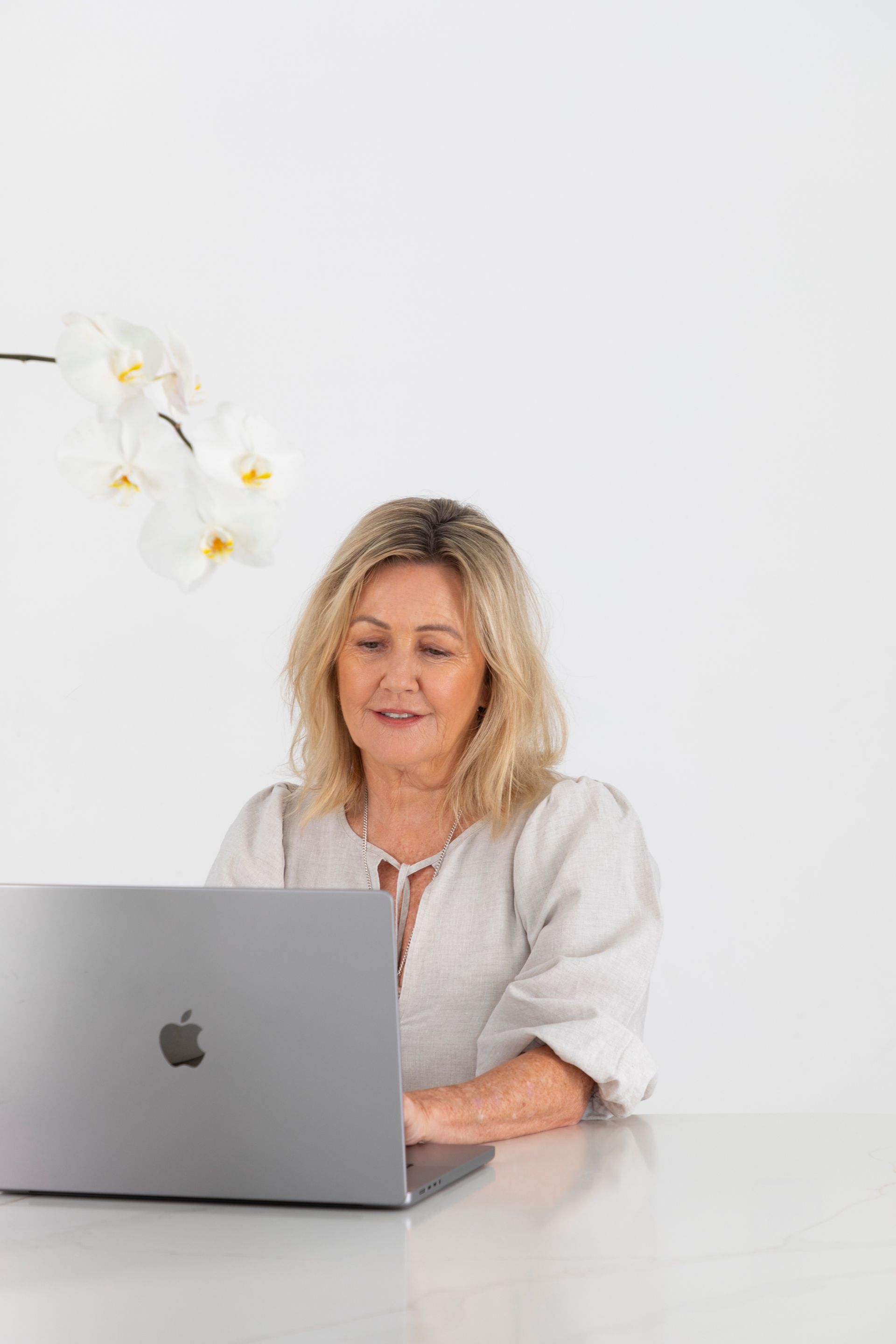 Woman with blonde hair working on a laptop, white table, white background, orchid flowers.
