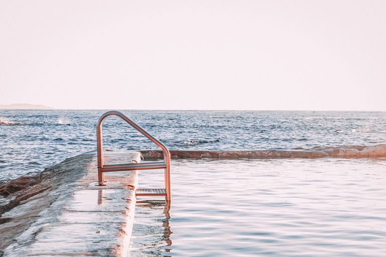 Sea pool with rusted ladder, concrete edge, and ocean view under a pale sky.