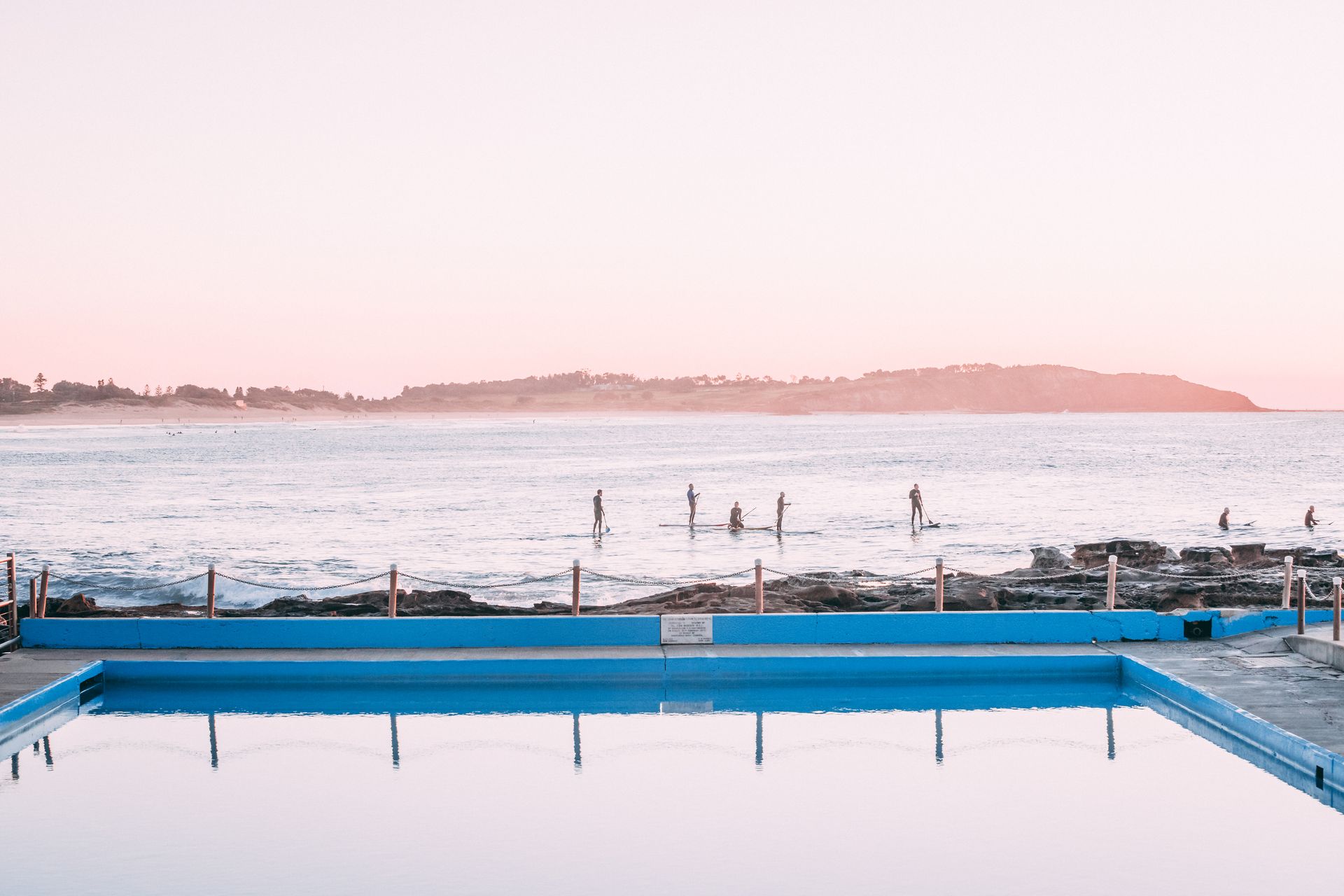 Blue ocean pool reflecting a pink sky and ocean with surfers.
