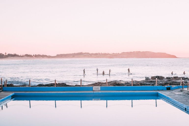 Blue ocean pool reflecting a pink sky and ocean with surfers.