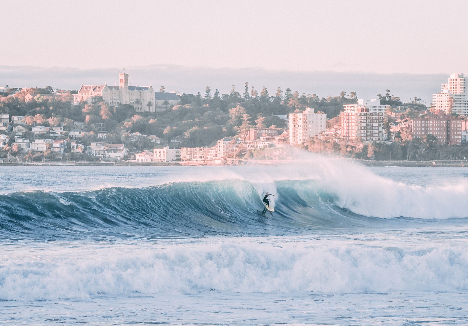 Surfer riding a wave near a city coastline. Ocean is turquoise.