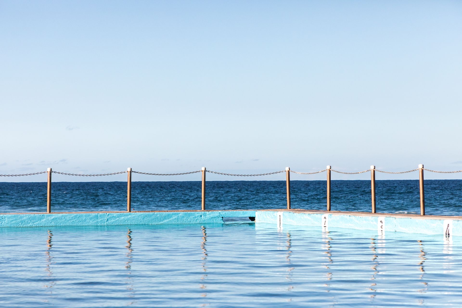 Ocean pool with blue water, fence, and ocean backdrop. Blue sky overhead.