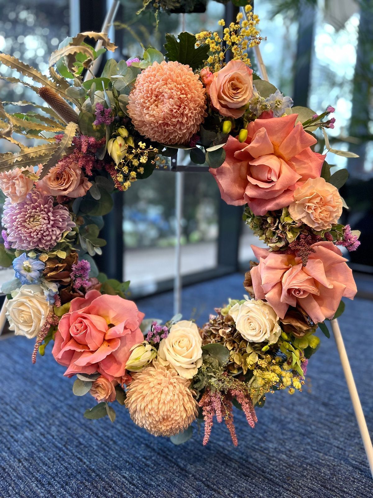 Floral wreath with coral roses, beige pom-poms, and greenery on a blue background.