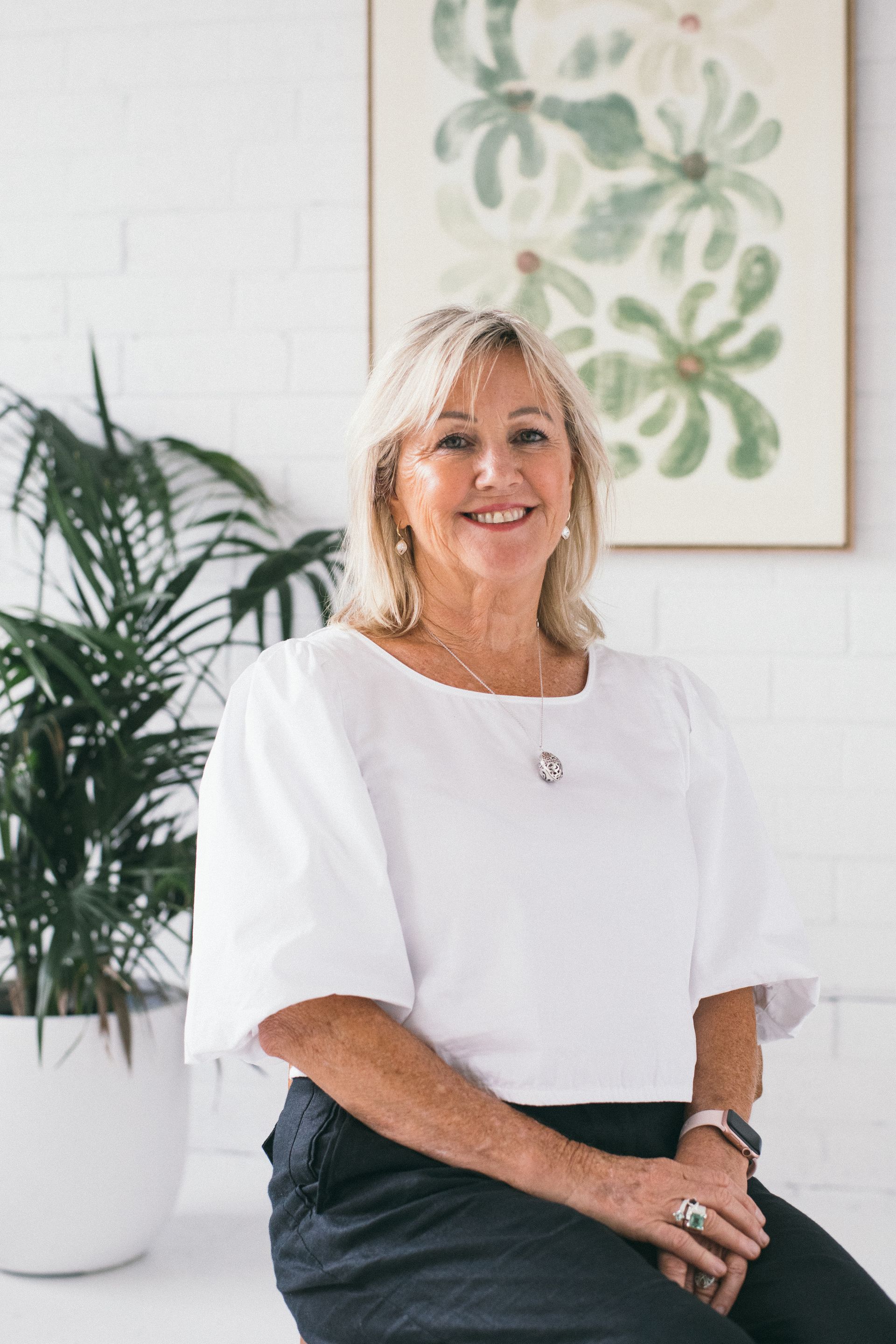 Woman in white top smiling, sitting near plant and artwork on white wall.