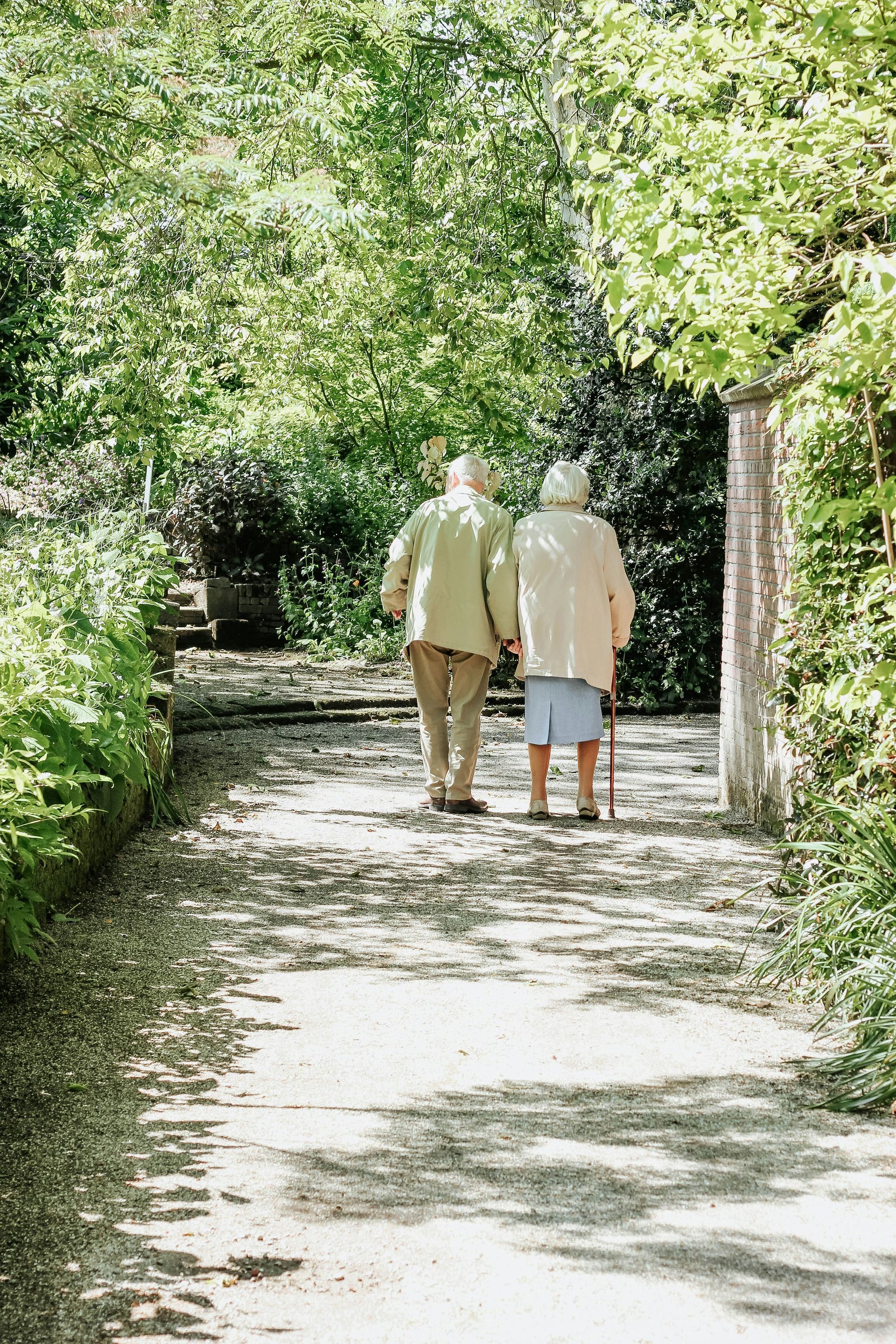 Elderly couple walking down a path, holding hands, surrounded by greenery.