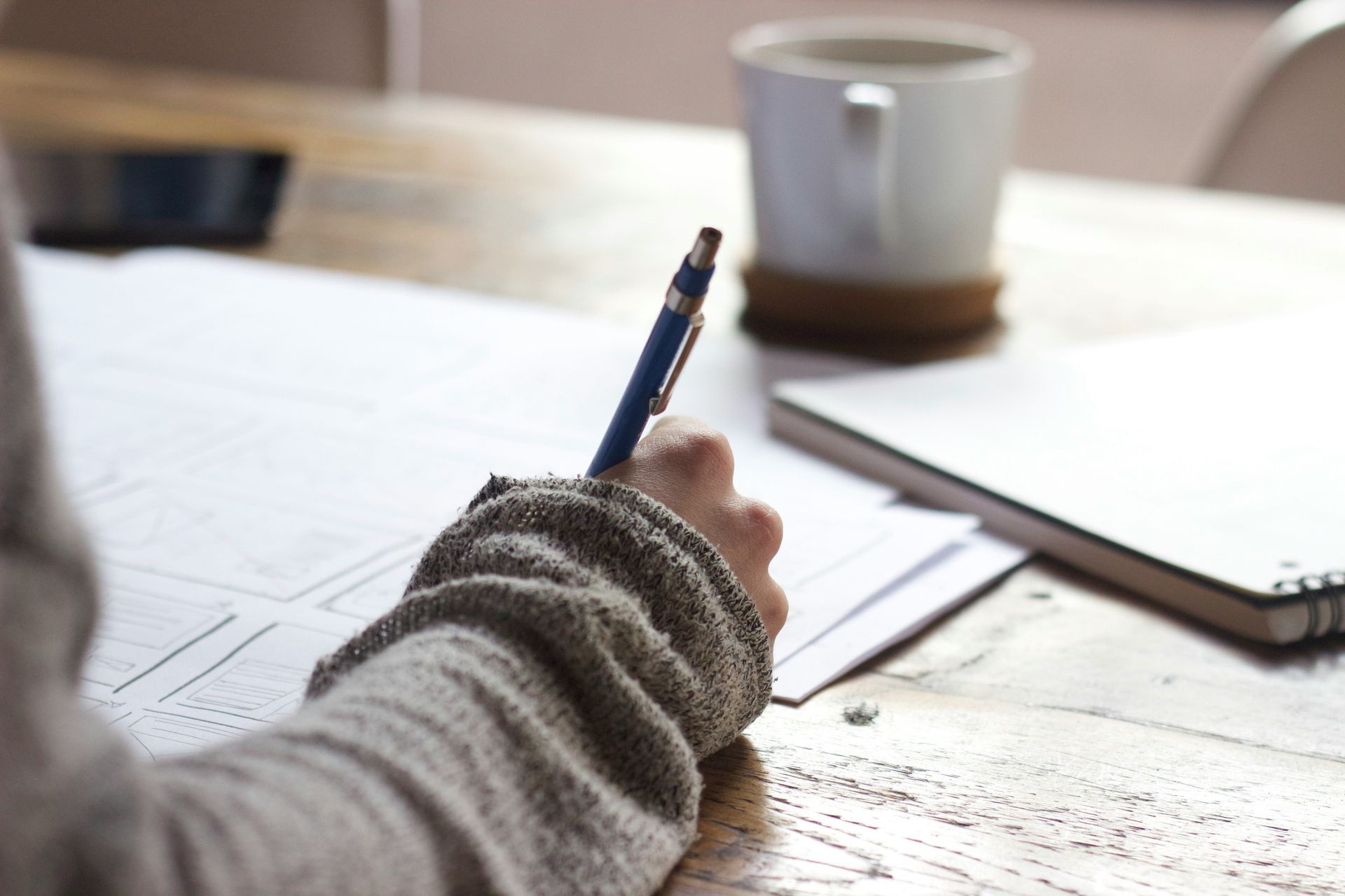 Person's hand writing with a pen on paper at a wooden table; a mug and notebook are in the background.