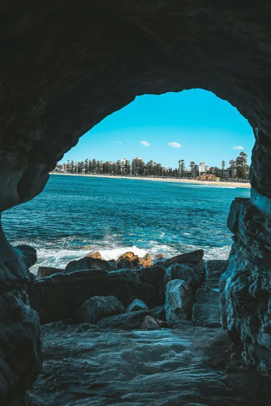 Ocean view through a rock cave opening. Blue water meets sandy beach, under a bright blue sky.