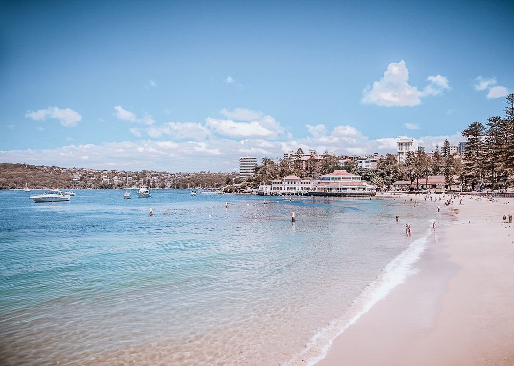 Beach with blue water, sandy shore, boats, buildings, and people under a blue sky.