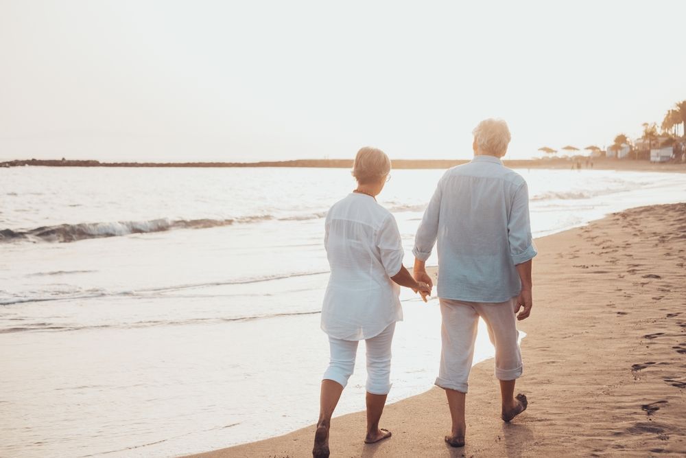 Elderly couple, holding hands, walking on beach at sunset.