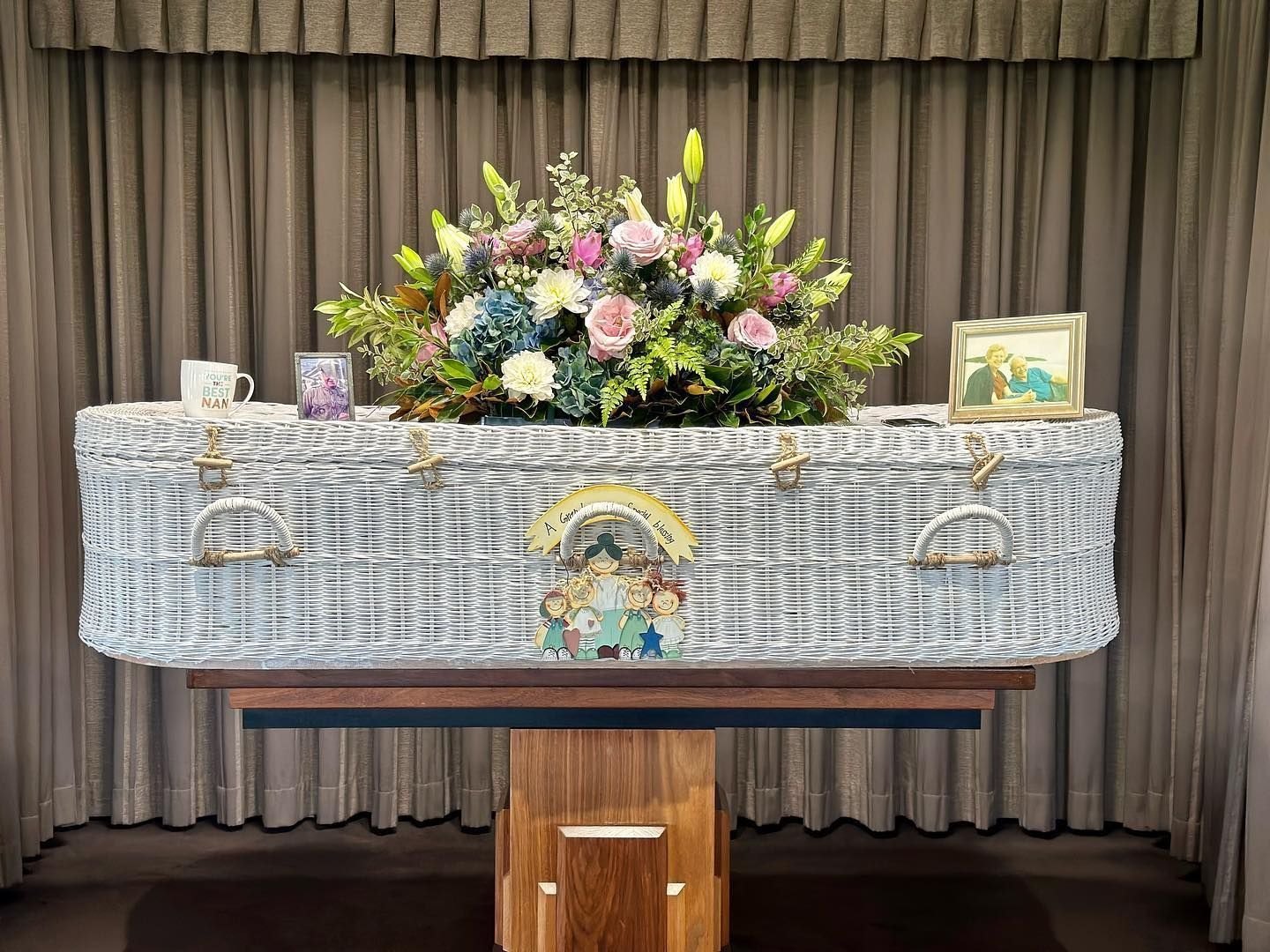 A casket draped with floral arrangements, set on a wooden stand, against a curtain backdrop.