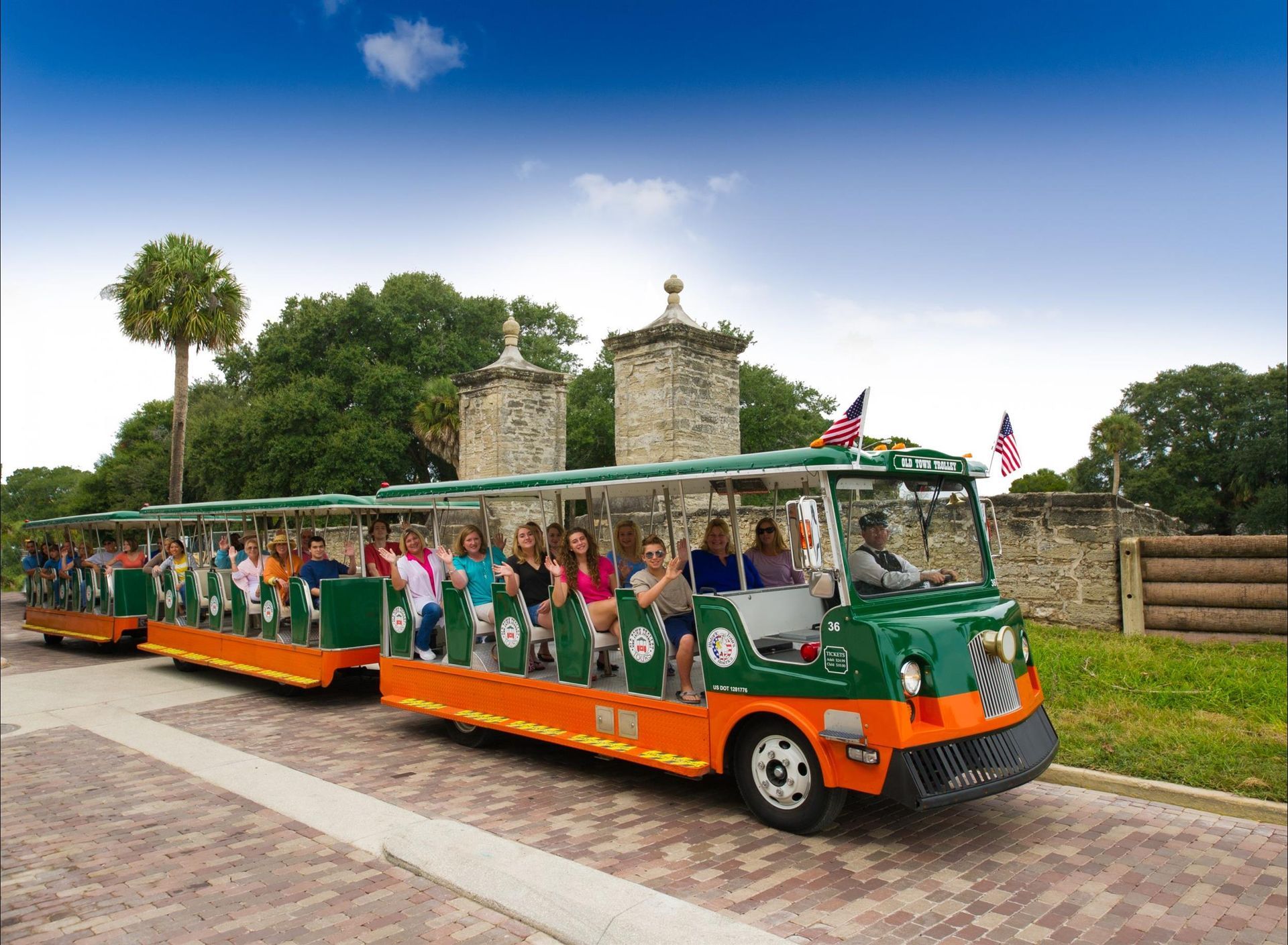 A green and orange bus filled with people is driving down a brick road.