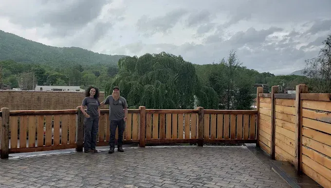 Two people standing on a patio with wooden fence, mountain backdrop. Overcast sky.