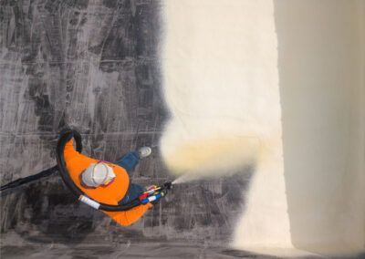 Person in orange vest spraying a wall with a light-colored coating.