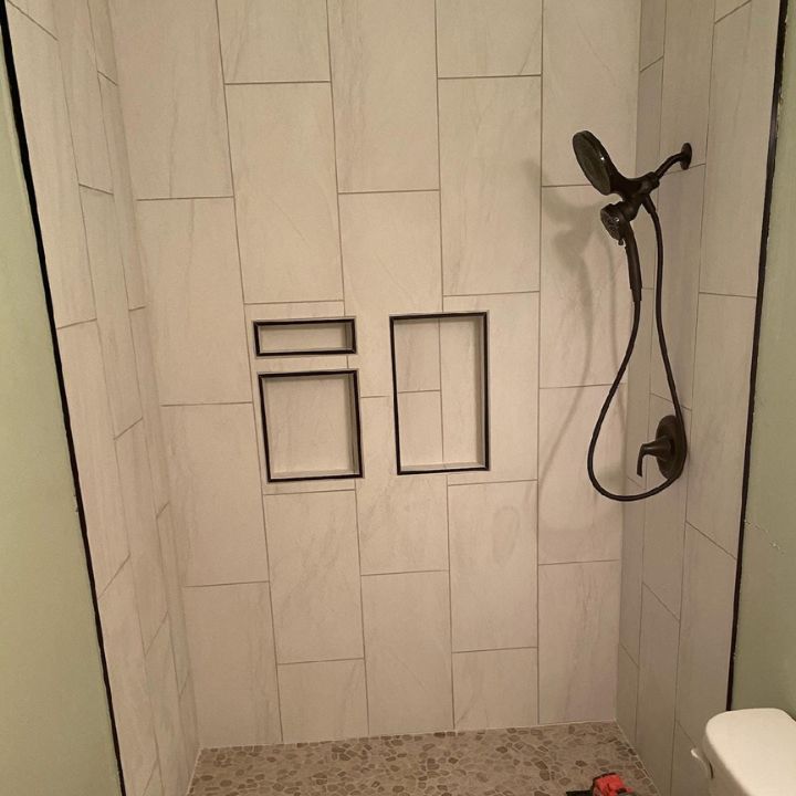 A modern shower with white marble-look tiles, dark recessed wall niches, and a dark handheld shower head.