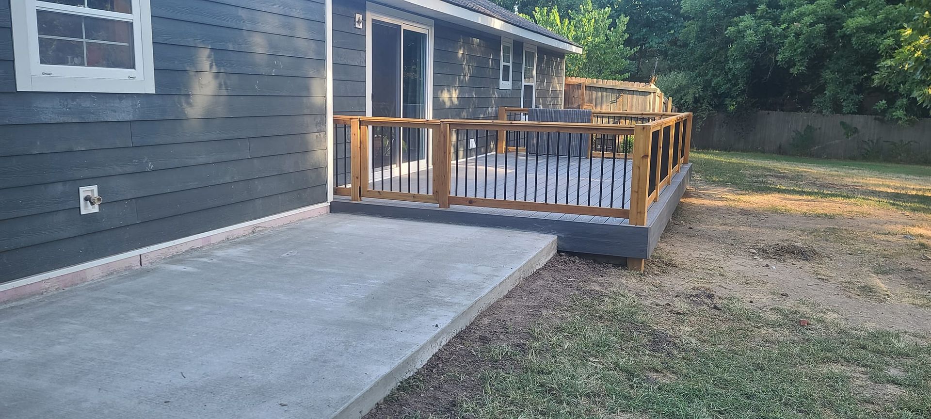 A gray house with a concrete patio leading to an elevated wooden deck with black railings in a backyard.