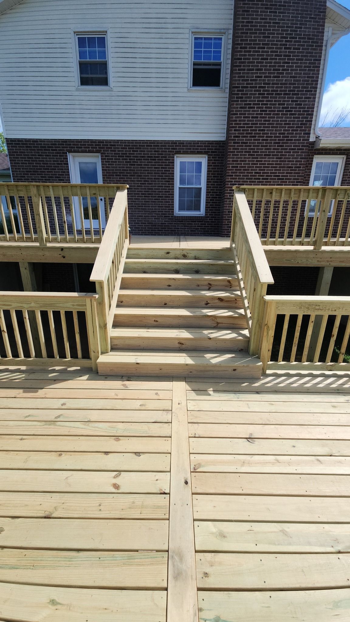 A wide, wooden deck with stairs leading up to the exterior wall of a two-story house with white siding and brick trim.