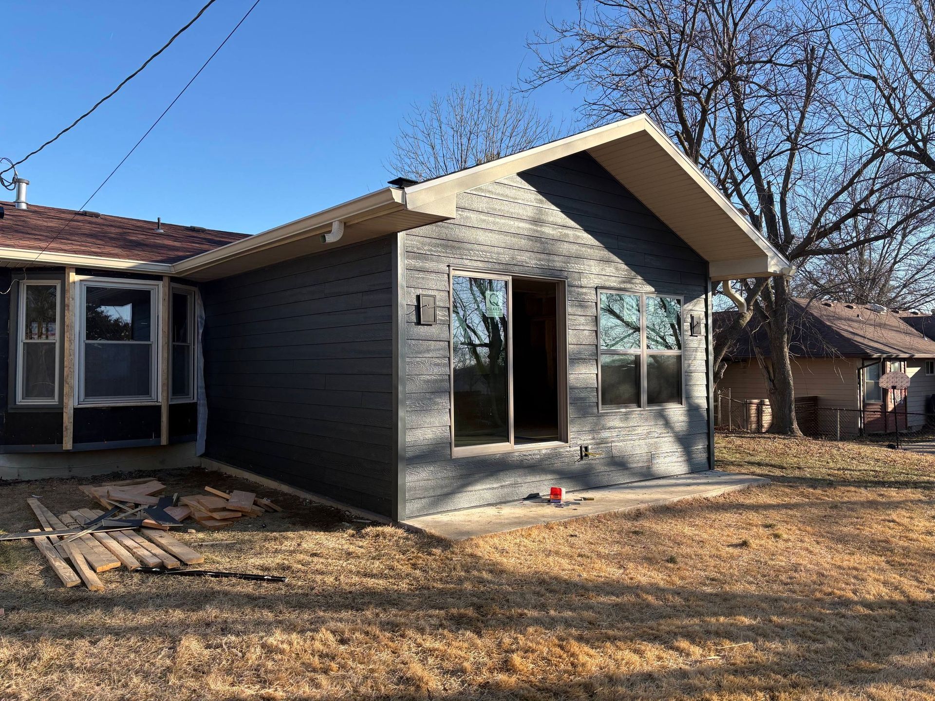 A house exterior renovation in progress, featuring newly installed gray stone siding and dark paneling under a blue sky.