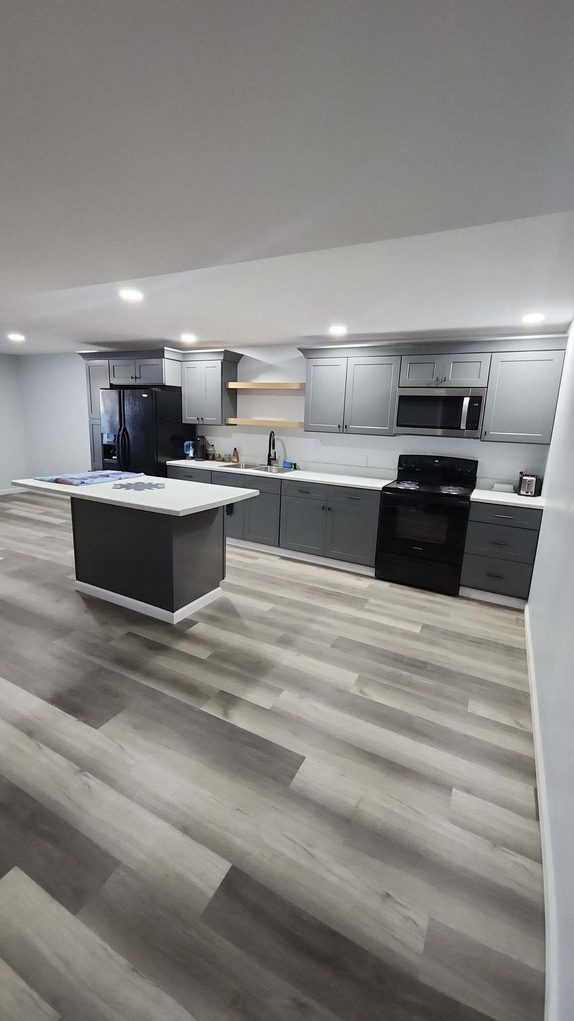 A modern, well-lit kitchen with gray cabinets, a central island, stainless appliances, and light wood plank flooring.