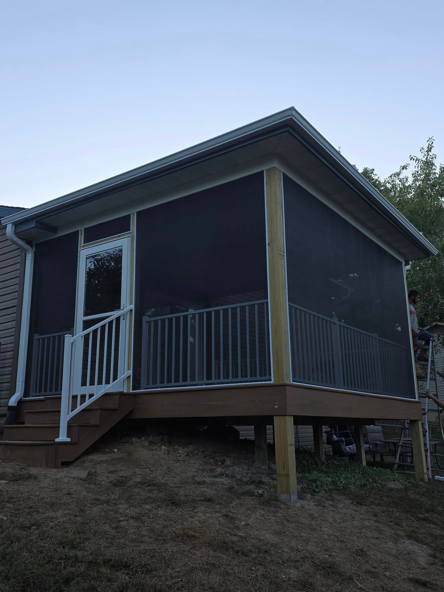 A wooden screened-in porch with a white railing and door, elevated on posts on a grassy slope under a clear sky.