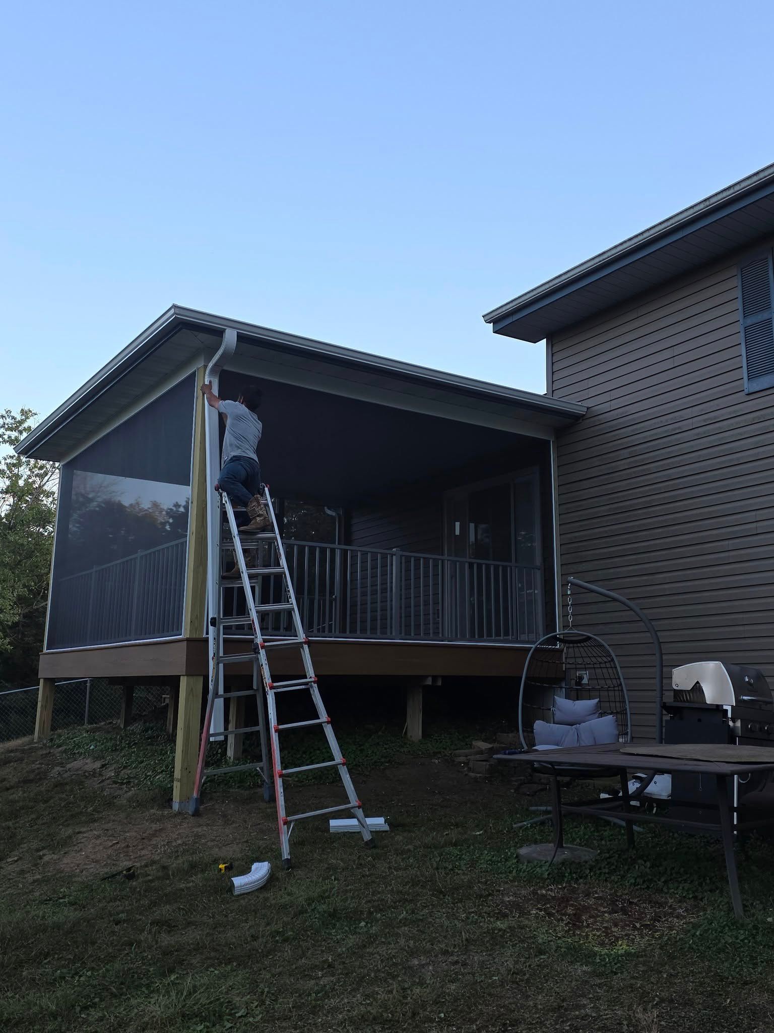 A person stands on a tall ladder, working on the screen enclosure of a raised back porch attached to a tan-colored house.