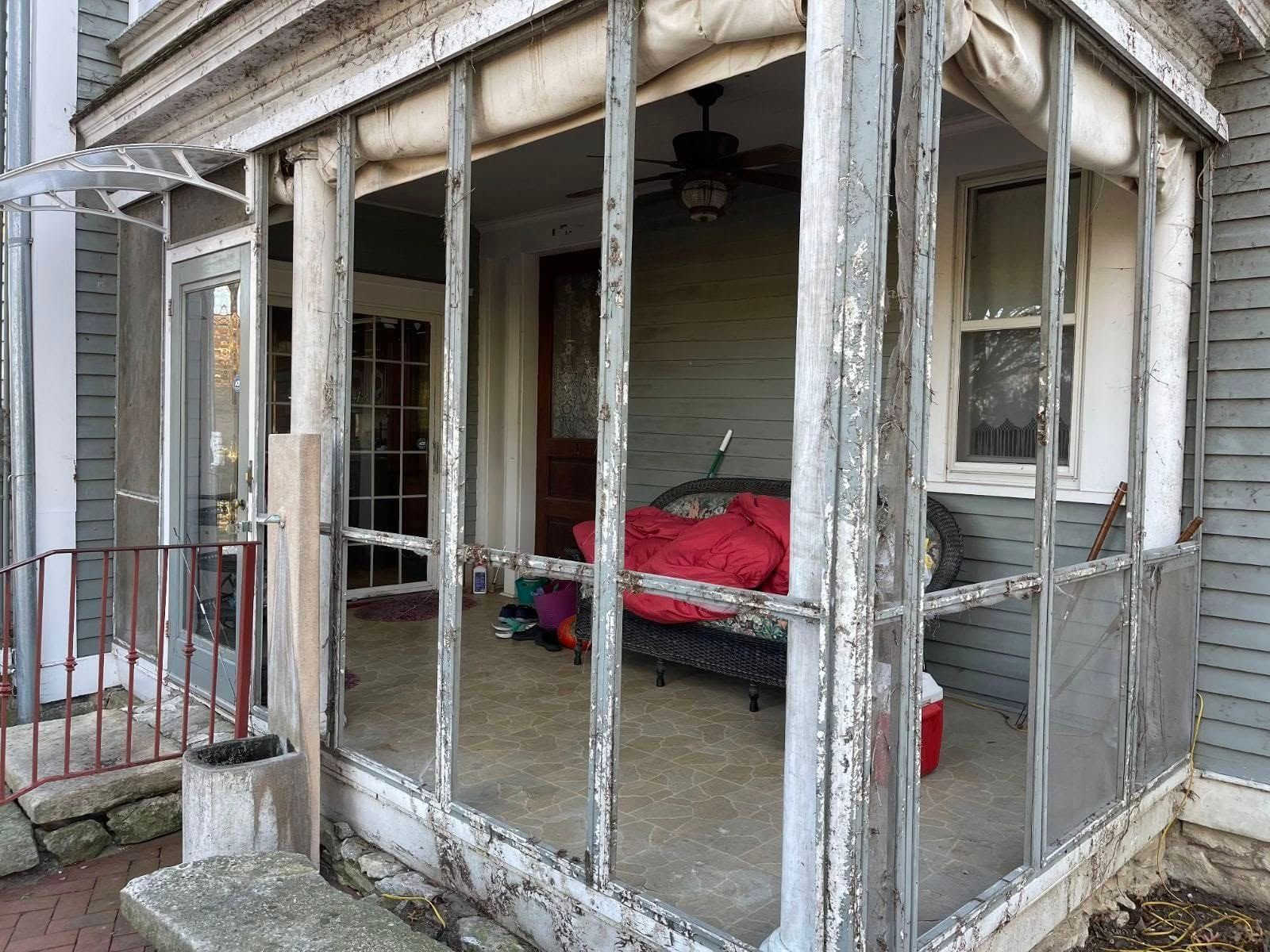 A weathered, enclosed porch with peeling gray paint, a red cushion on a bench, a ceiling fan, and a view into a house.